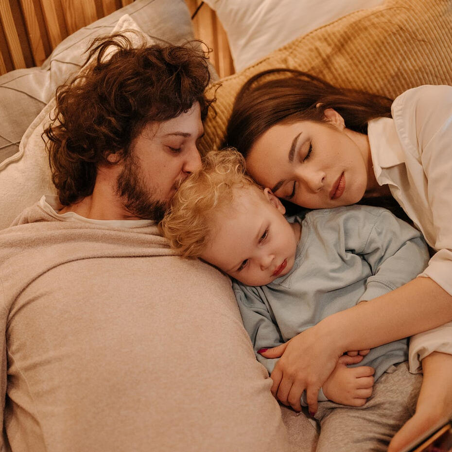 Mother and father asleep on a bed beside their baby, representing peaceful family sleep.
